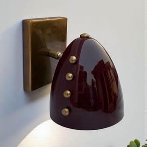 Op de afbeelding: Een wandlamp met een bordeauxrode kap en messing accenten. De glanzende kap heeft een druppelvorm met drie messing klinknagels. De lamp heeft een rechthoekige messing basis en een kleine messing arm.