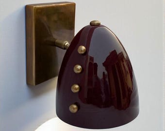 May include: A wall-mounted lamp featuring a glossy, deep burgundy, dome-shaped shade. The fixture includes a rectangular brass backplate and a brass arm connecting to the shade. Four small, round brass decorative studs are aligned on the side.