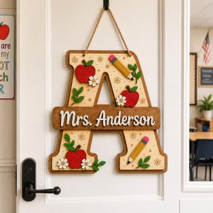 May include: A wooden letter A door sign featuring a natural wood finish with a horizontal bar displaying the white text Mrs. Anderson. The letter is decorated with red apples, yellow pencils, green leaves, and white flowers, suspended by a rope.