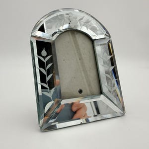 May include: An arched glass picture frame with beveled edges and a clear mirrored surface. The frame features etched floral and leaf patterns along the top arch and down both side panels, surrounding a central rectangular opening for a photo.