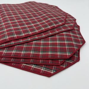 May include: A set of four hexagonal placemats featuring a red, dark green, and white plaid pattern. Each placemat has a solid red fabric border along the edges, creating a consistent look for a table setting with a classic textile design.