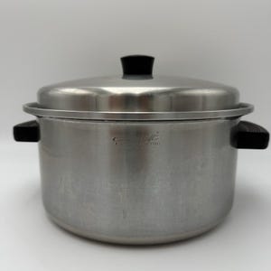 May include: A stainless steel stockpot with a matching lid, featuring black heat-resistant side handles and a black knob on the lid. The side of the pot is engraved with the text Regal Ware and Stainless Steel in a centered, horizontal orientation.