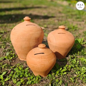 Könnte beinhalten: Drei Spardosen aus Terrakotta in verschiedenen Größen werden gezeigt. Jede Bank hat einen Schlitz für Münzen und eine abgerundete Form mit einem kleinen, dekorativen Aufsatz. Die Banken haben einen warmen, erdigen Ton und stehen auf grünem Gras.