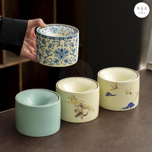 May include: A collection of ceramic containers with a cylindrical shape. One container is held by a person. The containers have various designs, including floral patterns, a cat with flowers, and flying birds. The containers are in shades of blue, green, and cream.
