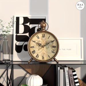 May include: A round, bronze-toned metal table clock featuring a vintage-style world map on its face. The clock has black Roman numerals, ornate black hands, and a circular top handle. It stands on three small, sturdy metal feet.