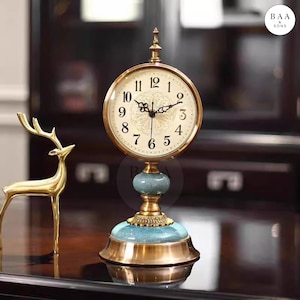 May include: A decorative table clock with a gold-coloured frame and a cream-coloured clock face. The clock has black numerals and hands. The base is teal and gold. A gold-coloured deer statue is to the left of the clock.