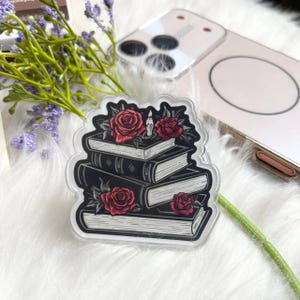 Op de afbeelding: Transparante acryl telefoonhouder met een ontwerp van zwarte boeken, rode rozen en een kaars. Het gotische ontwerp ligt op een witte, zachte ondergrond.