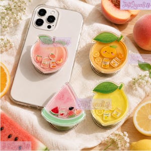 May include: Four fruit-themed phone grips featuring peach, orange, watermelon, and lemon designs. Each grip displays miniature icons of the corresponding fruit, juice cartons, and jars. One grip is attached to a smartphone, while three are separate.