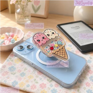 May include: A light blue smartphone case with a MagSafe ring and a clear, liquid-filled phone grip shaped like an ice cream cone. The grip features two smiling faces, a cherry, a heart, a star, and colorful sprinkles inside the clear casing.