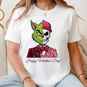 May include: White t-shirt featuring a split design of a green Grinch and a skull wearing a red hat. The Grinch side has a mischievous grin, while the skull side is stern. The shirt includes the text "Happy Valentine's Day!"