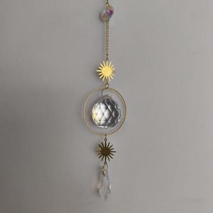 Puede incluir: Un atrapasoles decorativo con una cadena y detalles dorados. El atrapasoles presenta una bola de cristal facetada, un cristal transparente en forma de lágrima y dijes dorados en forma de estrella. Una pequeña cuenta de cristal iridiscente en la parte superior.
