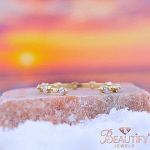 Può includere: Un delicato anello d'oro con piccole gemme trasparenti incastonate lungo la fascia. L'anello è esposto su una superficie strutturata e chiara con uno sfondo sfocato del tramonto. La parola "BEAUTIFY" è visibile in basso a destra.