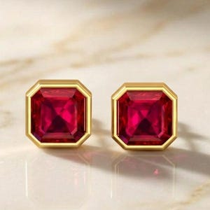 Può includere: Un paio di orecchini a lobo con pietre preziose rosso vivo dal taglio quadrato, incastonate in montature di metallo color oro lucido. Gli orecchini presentano una superficie sfaccettata che riflette la luce, creando un aspetto cremisi.