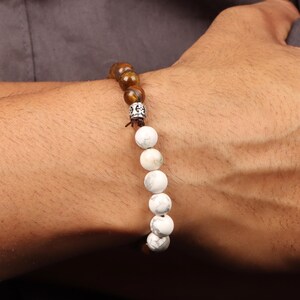 May include: A beaded bracelet featuring a combination of brown and white beads. The bracelet has a mix of round, tiger-eye-like beads and white, marbled beads. A silver-colored spacer bead is also included in the design. The bracelet is worn on a wrist.