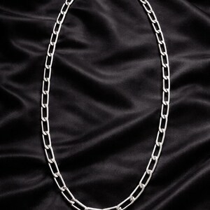 Puede incluir: Un collar de cadena plateada con un diseño de eslabones rectangulares. El collar se muestra sobre un fondo de satén negro, creando un contraste que resalta el brillo metálico de la joya. Es un accesorio sencillo y elegante.
