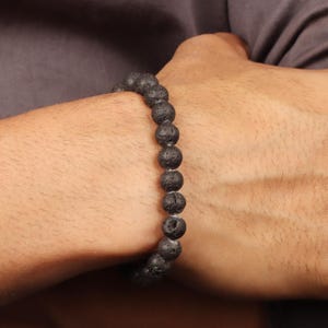 May include: A black lava rock bead bracelet. The bracelet is made up of a series of round, porous beads. The beads are a dark, charcoal colour, and the bracelet is worn on the wrist.