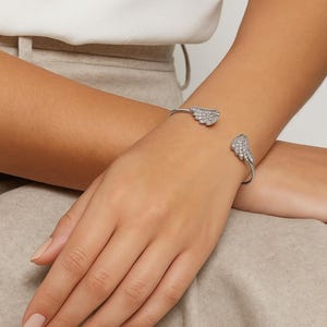 Puede incluir: Un brazalete plateado con dise&ntilde;os de alas adornados con peque&ntilde;os cristales brillantes. El brazalete abierto se muestra en una mu&ntilde;eca, mostrando los detalles delicados de la joya.
