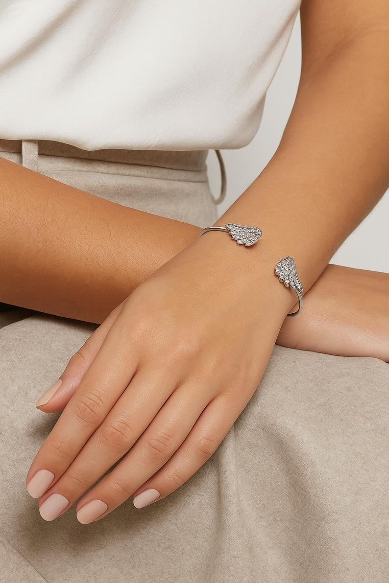 Puede incluir: Un brazalete plateado con dise&ntilde;os de alas adornados con peque&ntilde;os cristales brillantes. El brazalete abierto se muestra en una mu&ntilde;eca, mostrando los detalles delicados de la joya.