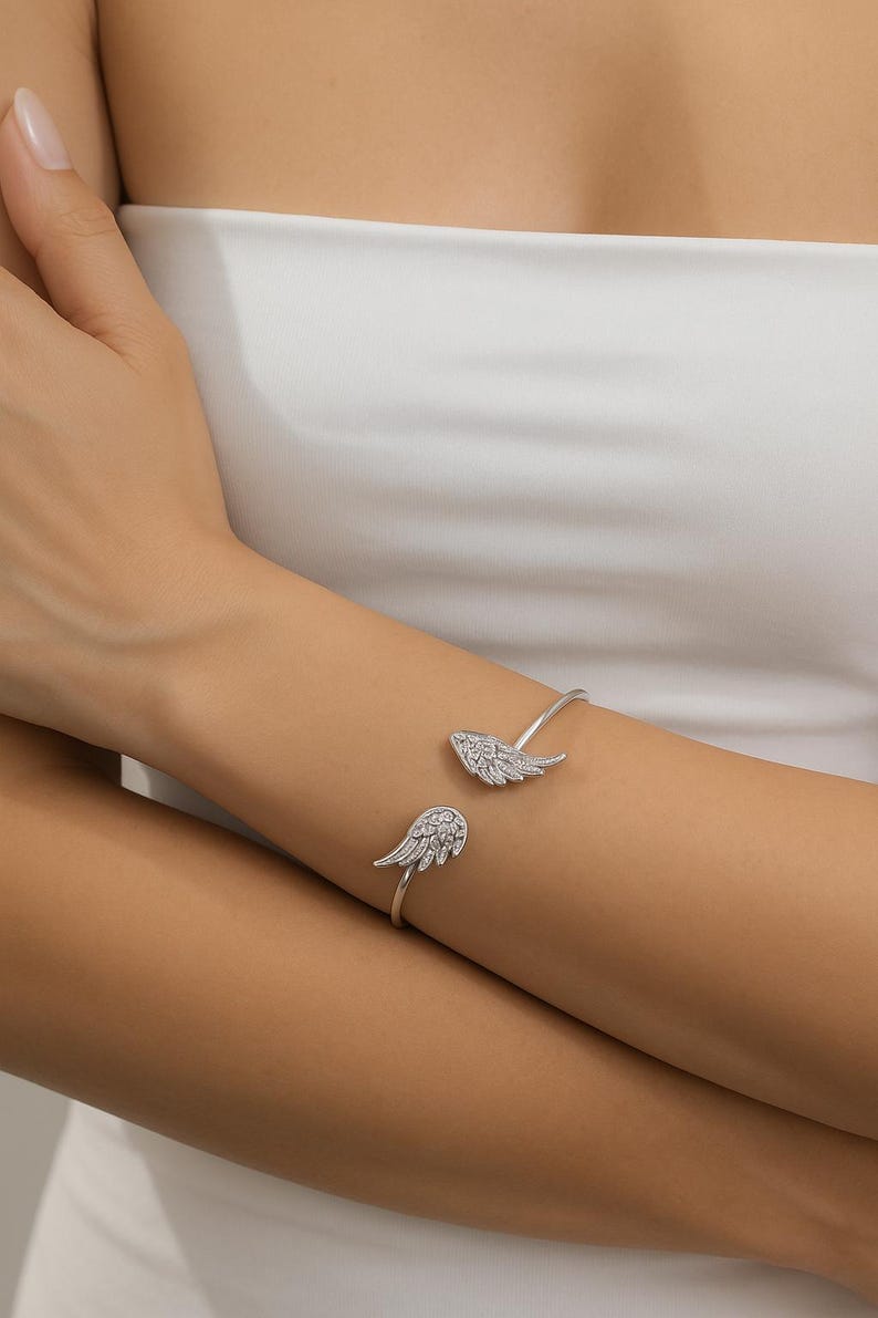 Puede incluir: Una pulsera de pu&ntilde;o plateada con dos dise&ntilde;os en forma de alas, cada uno adornado con peque&ntilde;os cristales brillantes. La pulsera se lleva en una mu&ntilde;eca, mostrando su est&eacute;tica delicada y elegante. Esta joya es un accesorio elegante.