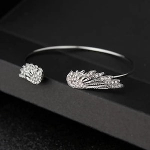Puede incluir: Un brazalete de pu&ntilde;o plateado con un dise&ntilde;o de ala adornado con cristales transparentes. El brazalete se muestra sobre una superficie negra.