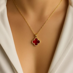 Puede incluir: Collar dorado con un colgante de trébol de cuatro hojas con centro rojo vibrante. El trébol está delineado en dorado y cuelga de una delicada cadena. El collar se luce sobre una camisa blanca.