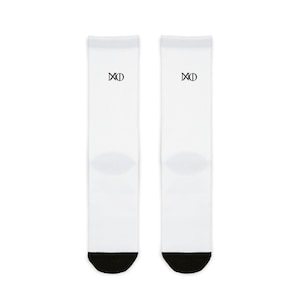 May include: A pair of white crew socks featuring a black heel and toe section. Each sock displays a small, centered black graphic logo consisting of a stylized letter M and a circle symbol positioned on the upper portion of the white fabric.