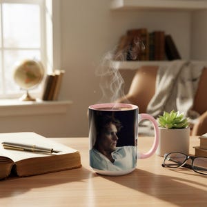May include: A white ceramic mug with a pink interior and handle, displaying a man's portrait. Steam rises from the dark liquid within. The mug is on a wooden table, beside an open book, a pen, and spectacles.