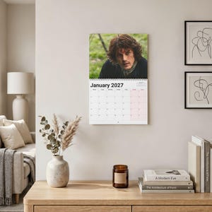 May include: A wall calendar for January 2027 featuring a photographic portrait of a person with wavy brown hair and a dark jacket at the top, with a grid of dates below, hanging on a light-colored wall above a wooden desk with decor.