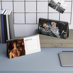 Może przedstawiać: Dwa spiralnie oprawione kalendarze biurkowe. Jeden kalendarz wyświetla miesiąc sierpień 2026 ze zdjęciem mężczyzny. Drugi kalendarz zatytułowany "Outlander 2026 Calendar" ze zdjęciem pary. Czarno-białe zdjęcie mostu jest przypięte do ściany.