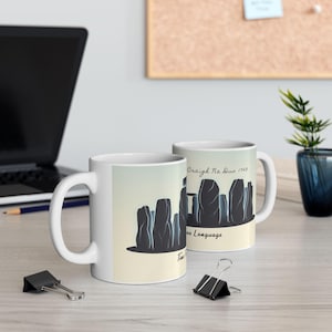 Puede incluir: Dos tazas de cerámica blanca con un gráfico de un círculo de piedra. El diseño presenta siluetas grises oscuras de piedras verticales sobre un fondo degradado amarillo claro y azul. El texto en las tazas dice "Craigh Na Dun 1743" y "Time is the Language".