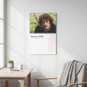 Può includere: Calendario da parete per gennaio 2026 con un ritratto di un uomo con i capelli castani. Il calendario è bianco con rilegatura a spirale e una griglia di date. Il calendario è appeso a una parete bianca.