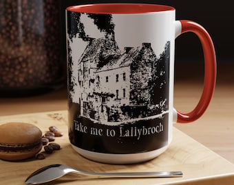 May include: White ceramic mug with a red interior and handle. The mug features a black and white illustration of a building and the text "Take me to Lallybroch". A macaron and spoon are on a wooden surface.