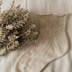 May include: A cream-colored knit sock featuring a delicate eyelet lace pattern throughout the leg and foot, finished with a subtle ruffled edge at the cuff. The sock is displayed alongside a cluster of dried, textured off-white flowers.