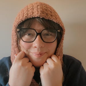 May include: A crocheted hood in a muted peach color. The hood is made of a textured yarn and has a close-fitting design. The person in the image is wearing black-framed glasses and a dark blue shirt.