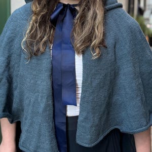 May include: A blue knitted cape with a hood and a dark blue ribbon. The cape has a flared shape and is made of a soft, textured material. The cape is worn over a white top.
