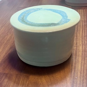 Peut inclure: Un beurrier en céramique cylindrique avec une finition émaillée blanc cassé lisse. Le couvercle présente un motif de cercle bleu clair texturé peint à la main sur sa surface supérieure. La base du récipient est légèrement plus large.