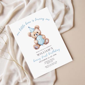 May include: A white invitation card with a watercolor illustration of a teddy bear holding a blue balloon. The text reads "our little bear is turning one" and announces William's first birthday on October 25th at 3 PM.