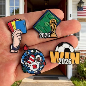 Puede incluir: Un conjunto de pines de esmalte con temática de fútbol. Un pin muestra una mano sosteniendo tarjetas rojas, amarillas y azules. Otros pines incluyen un campo de fútbol con un trofeo y el año 2026, un balón de fútbol con la palabra "WIN" y el año 2026.