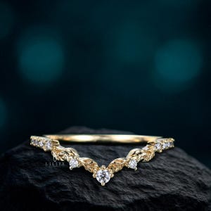 May include: A gold-colored ring with a leaf and diamond design. The ring has a curved shape with small diamonds set along the top and a larger diamond at the center. The ring is displayed on a dark, textured surface.