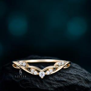 May include: A gold-colored ring with a curved design, featuring multiple small, clear gemstones. The ring has a delicate, elegant appearance, with the gemstones set along the curve. The ring is set against a dark background.