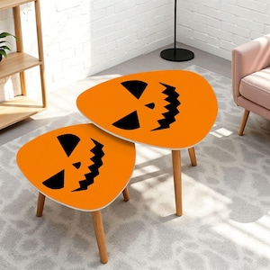 May include: A set of two nesting tables with orange, irregular, rounded tops featuring black, jagged, triangular eyes and a serrated mouth design. Each table is supported by three light-colored wooden legs, creating a functional furniture piece.