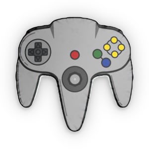 Könnte beinhalten: Ein graues Kissen in Form eines klassischen Videospiel-Controllers. Der Controller hat ein Steuerkreuz, eine zentrale graue Taste und farbige Tasten in Gelb, Grün und Blau. Das Design ist schwarz umrandet.