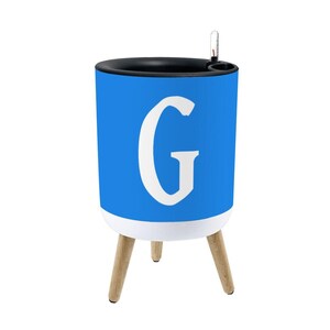 May include: A cylindrical blue planter featuring a large white letter G on the front. The planter has a black rim, a white base, and three light-colored wooden legs. A small clear water level indicator is attached to the top rim of the planter.