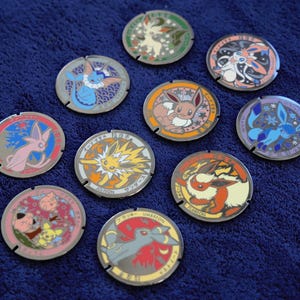 Puede incluir: Colección de fichas metálicas redondas y coloridas, cada una con un personaje Pokémon diferente. Las fichas presentan colores vibrantes y texto japonés. Están dispuestas sobre una superficie azul oscuro.
