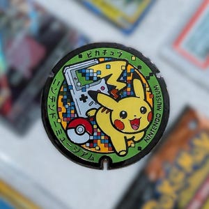 May include: A circular, green and black manhole cover featuring a yellow Pikachu character, a Game Boy, and a Poké Ball. The manhole cover has Japanese text and is surrounded by Pokémon trading cards.