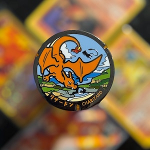 Puede incluir: Pin circular de esmalte con Charizard, una criatura naranja con forma de dragón, sobre un cielo azul y un paisaje verde. El pin incluye texto japonés y la palabra "CHARIZARD".
