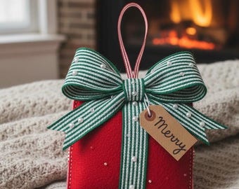 May include: A red felt Christmas ornament shaped like a gift box, adorned with a green and white striped bow and faux pearls. A small tag with the word "Merry" hangs from the bow. The ornament has a red and white striped hanging loop.