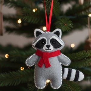 May include: A gray felt raccoon Christmas ornament with a red scarf and a black and white striped tail. The ornament has white stitching and a red ribbon for hanging. The raccoon has black eyes and a black nose.