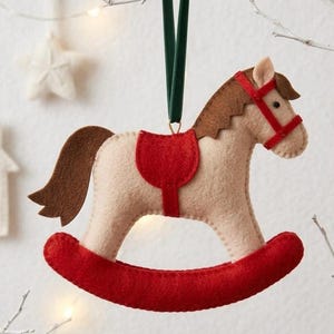 May include: A felt rocking horse Christmas ornament. The horse is cream-colored with a brown mane and tail, and a red saddle and bridle. The ornament hangs from a green ribbon and is set against a white background.