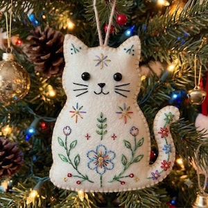 May include: A white felt cat ornament featuring black bead eyes and embroidered details. The cat is decorated with colorful floral and leaf patterns in blue, green, red, and yellow thread. It hangs from a simple twine loop for display.